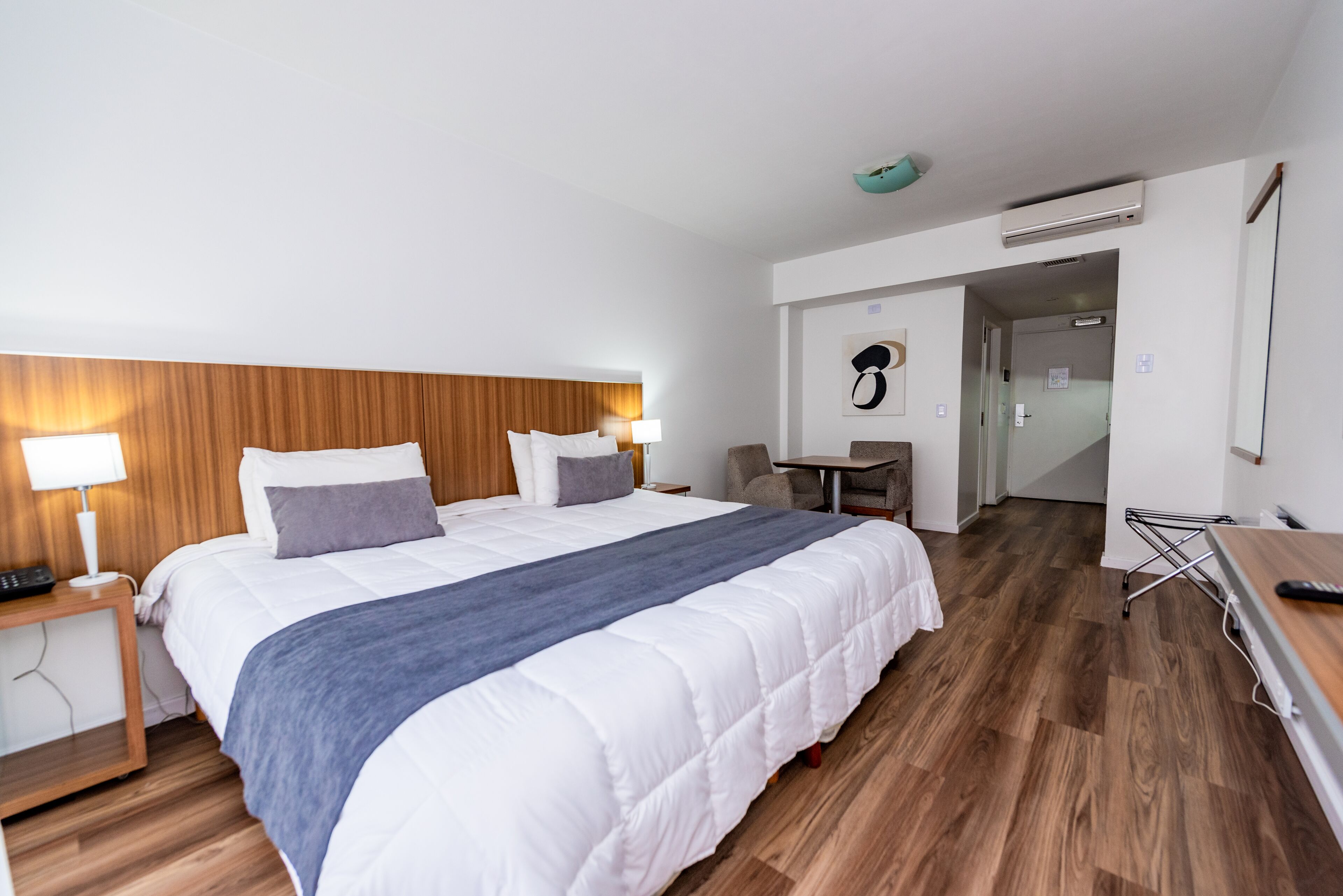 Habitación de hotel con una cama grande, cabecera de madera, mesitas de noche con lámparas, un escritorio y un equipo de aire acondicionado.