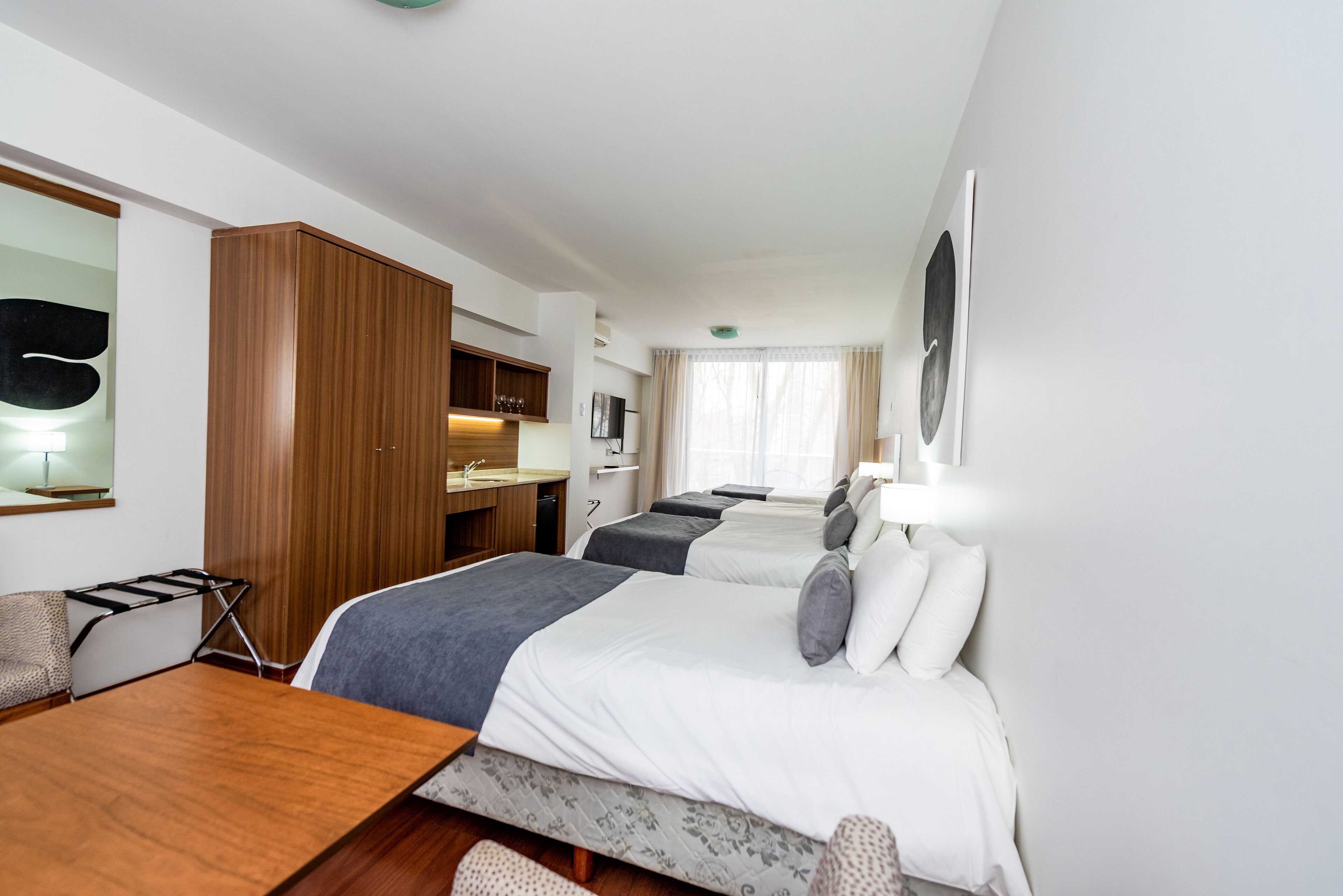 Quarto de hotel com duas camas, um guarda-roupa de madeira, uma mesa de trabalho e uma kitchenette pequena.