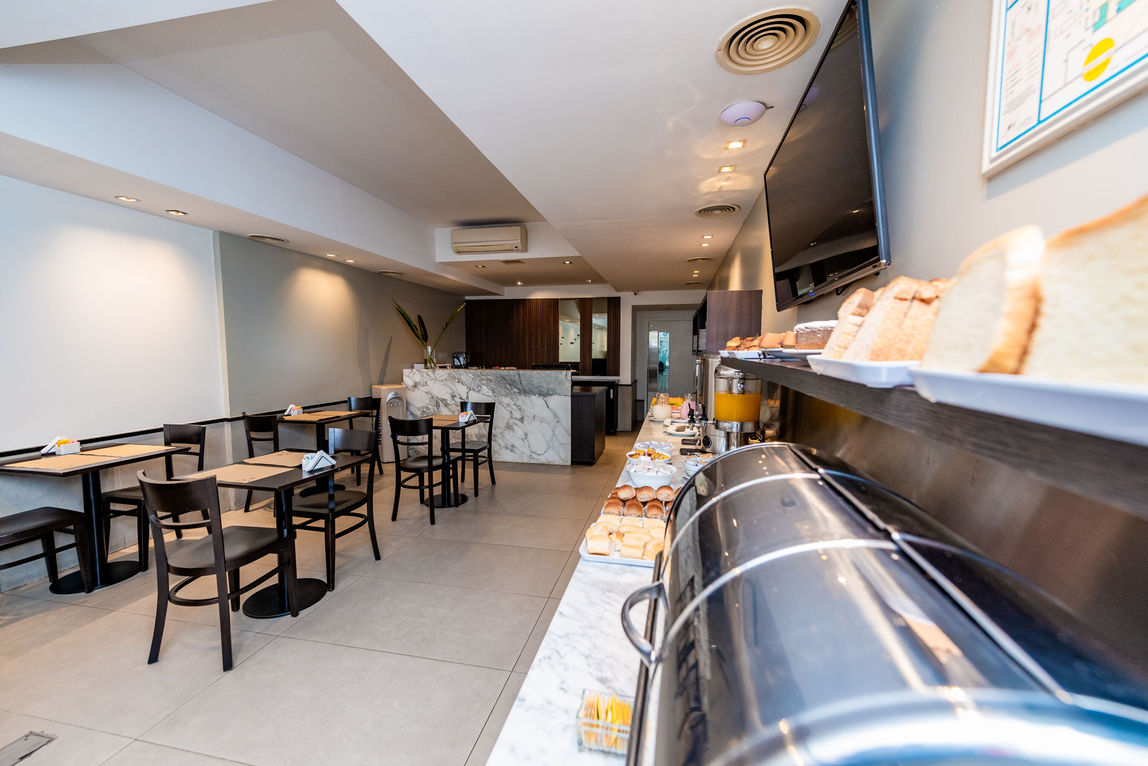A modern hotel dining area with marble countertops, buffet with bread, and a TV mounted on the wall.