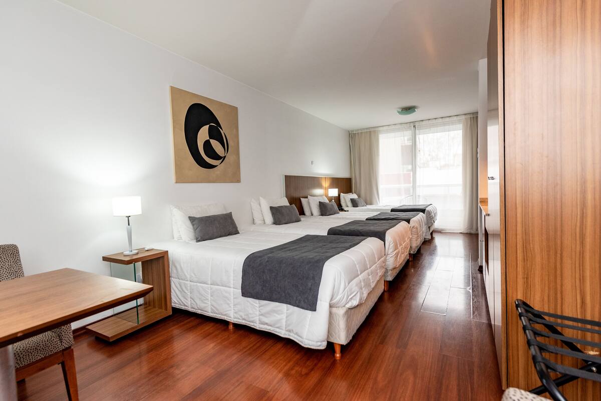 A hotel room with a long bed, a desk, a chair, and a large window.