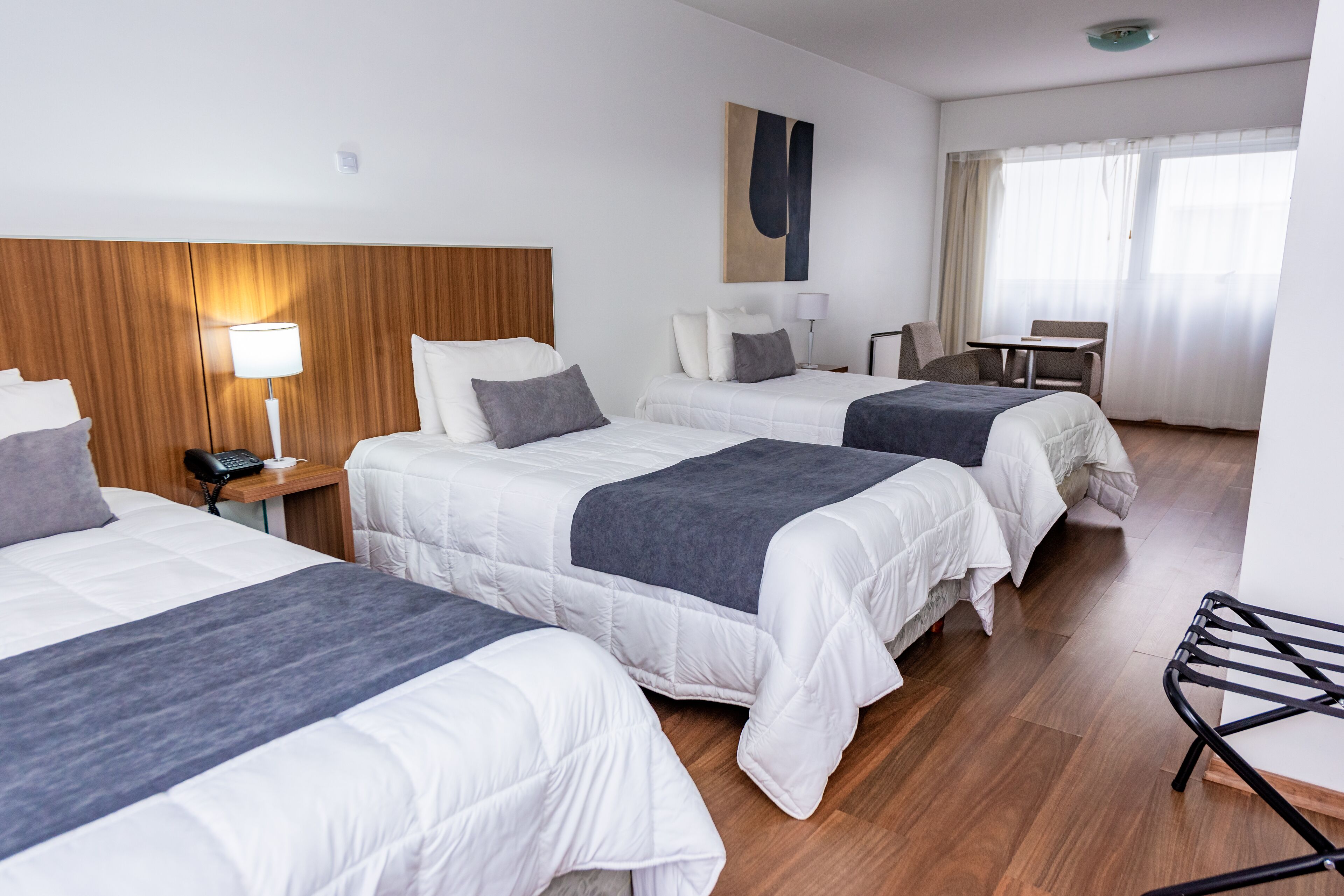 Una habitación de hotel con tres camas, un cabecero de madera, un escritorio con una silla y una ventana con cortinas.