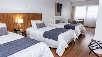 Ett hotellrum med tre sängar, en sänggavel i trä, ett skrivbord med en stol och ett fönster med gardiner.