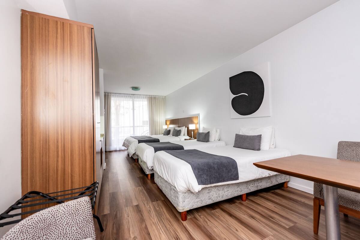 A hotel room with a large bed, a smaller bed, a desk, and a chair. There is a wardrobe and a window with curtains.