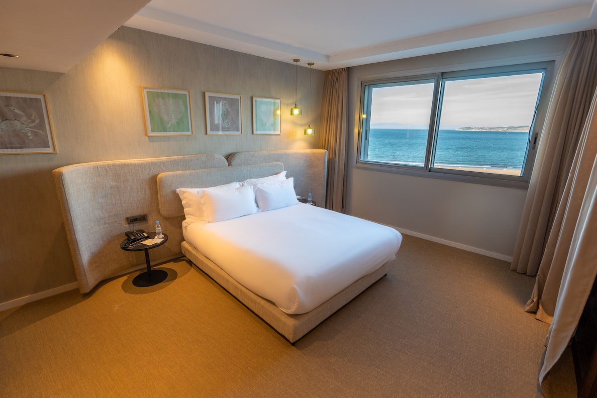A hotel room with a large bed, a small table with a bottle, and a view of the sea through a large window.