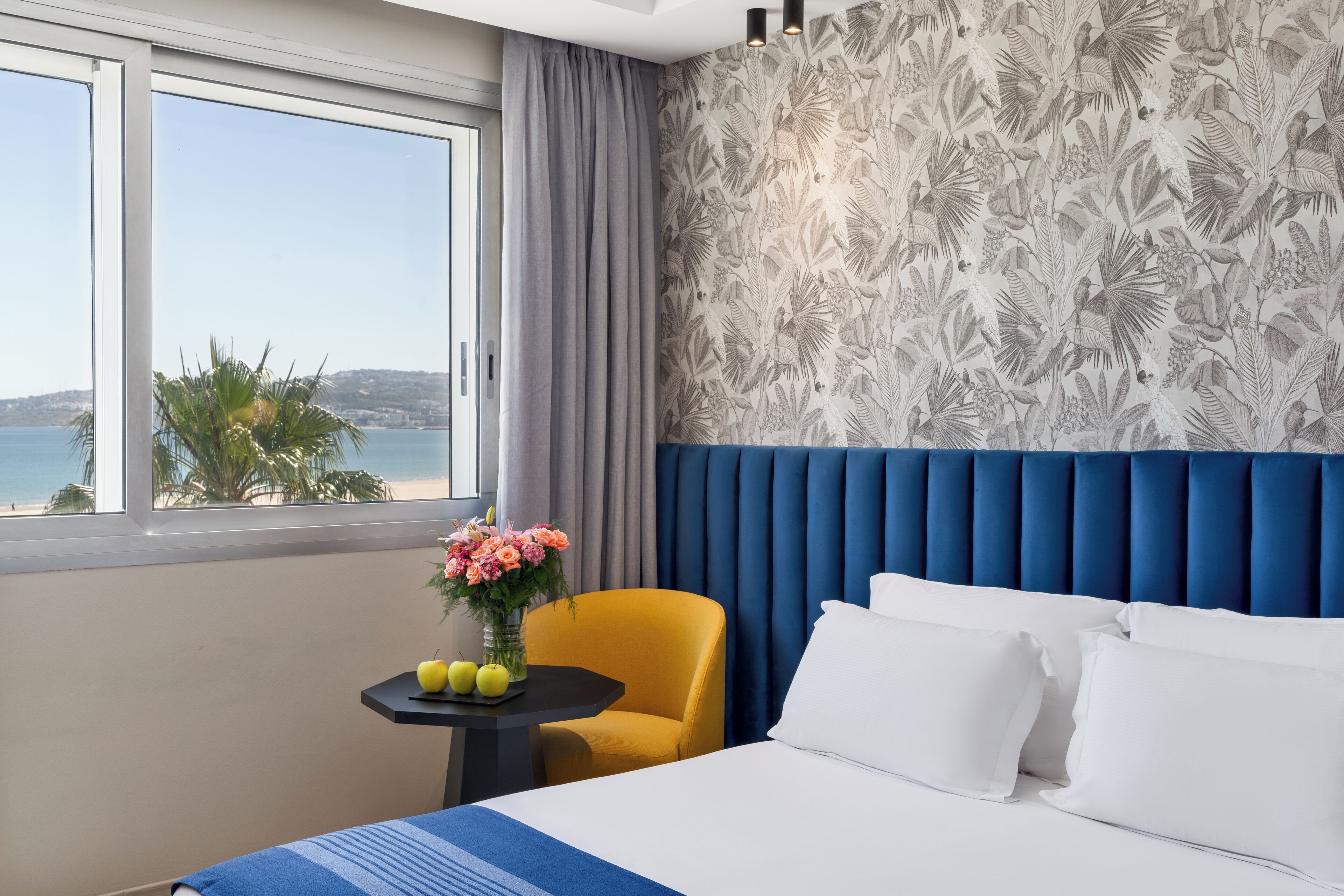 A hotel room with a large bed, a view of the sea, a tropical wallpaper, and a small table with fruit and flowers.