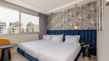 Habitación de hotel con una cama grande, una mesita y vistas de la ciudad a través de un ventanal amplio.