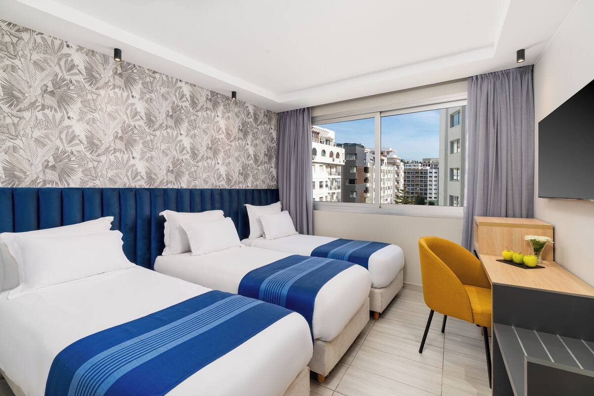 A hotel room with two beds, a desk, a chair, and a large window with a city view.