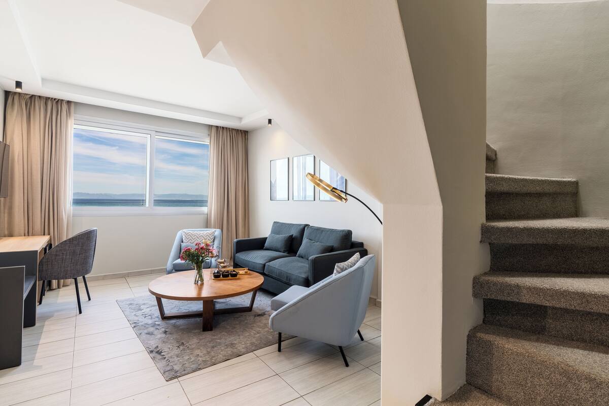 A modern living room with a sofa, armchair, coffee table, and a view of the sea through large windows.
