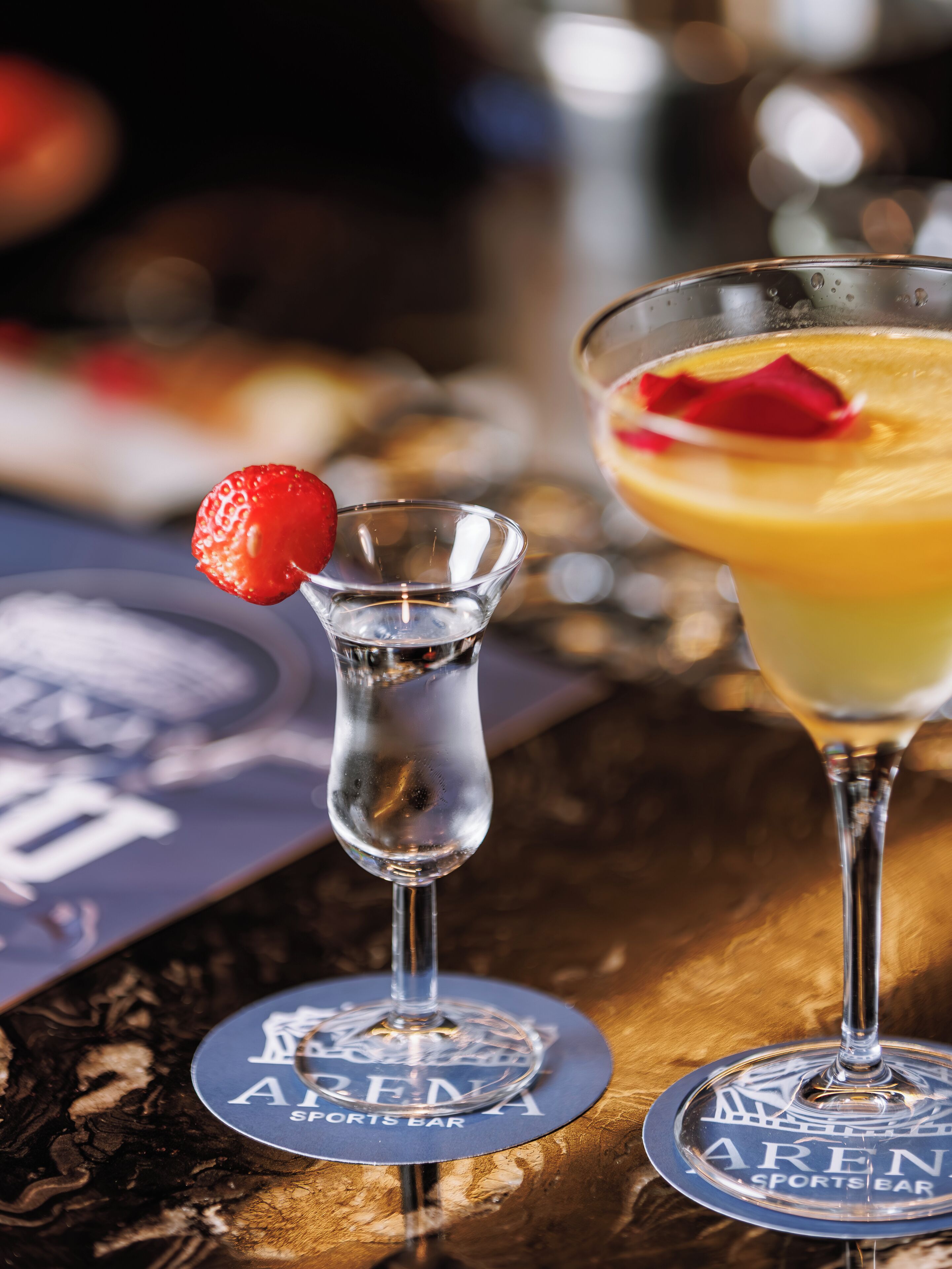 Two glasses of beverages with a strawberry garnish on one and a rose on the other, placed on a table with a coaster that reads "ARENA SPORTS BAR".