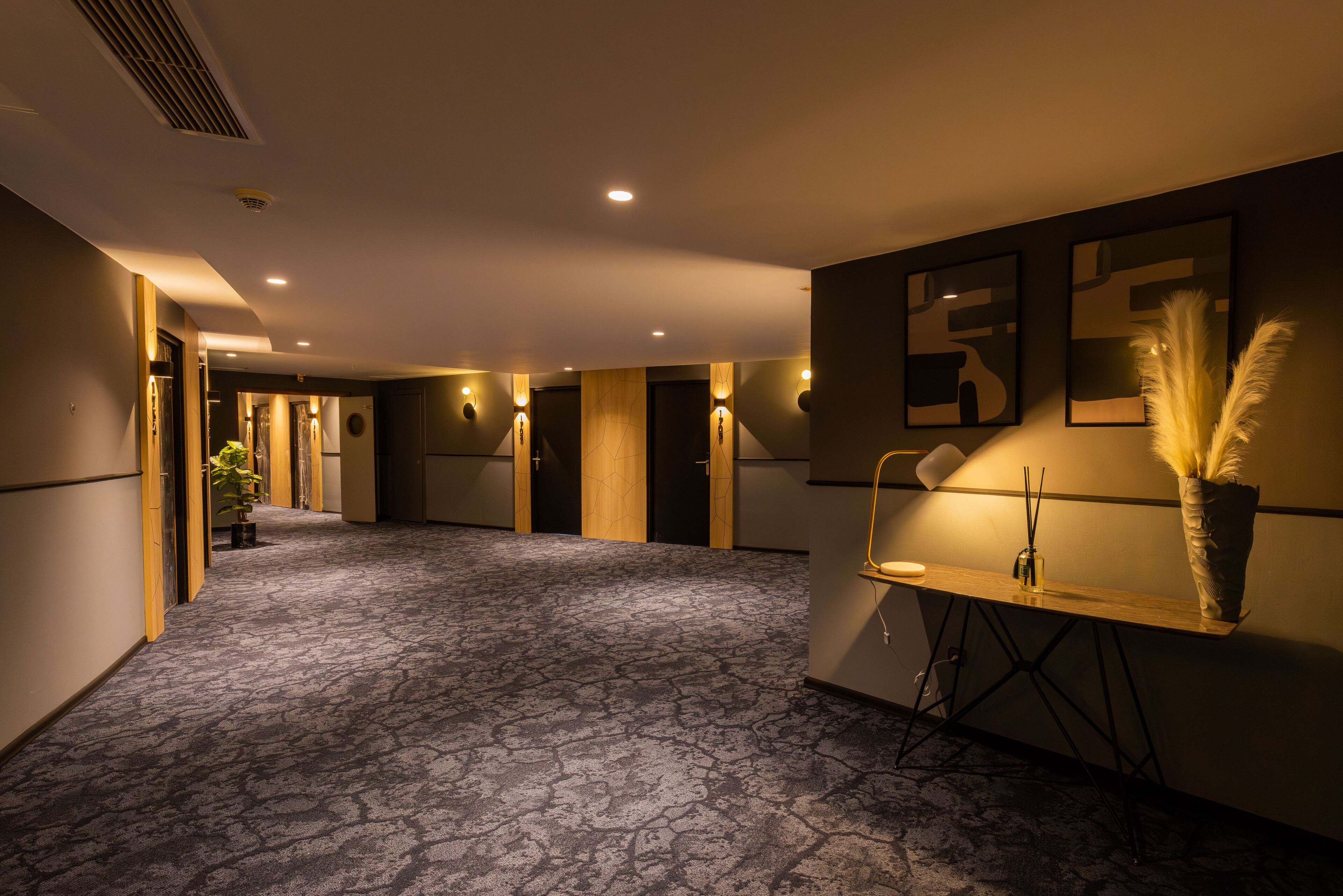 Een hotelgang met een kamerbreed tapijt, ingebouwde verlichting en kunstwerken in lijsten aan de wanden.