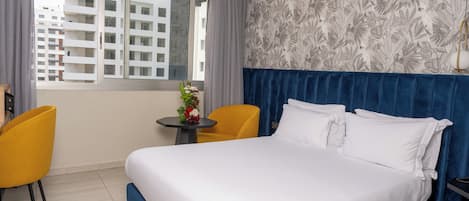 A hotel room with a large bed, two yellow chairs, a small table with a vase of flowers, and a window with a view of buildings.