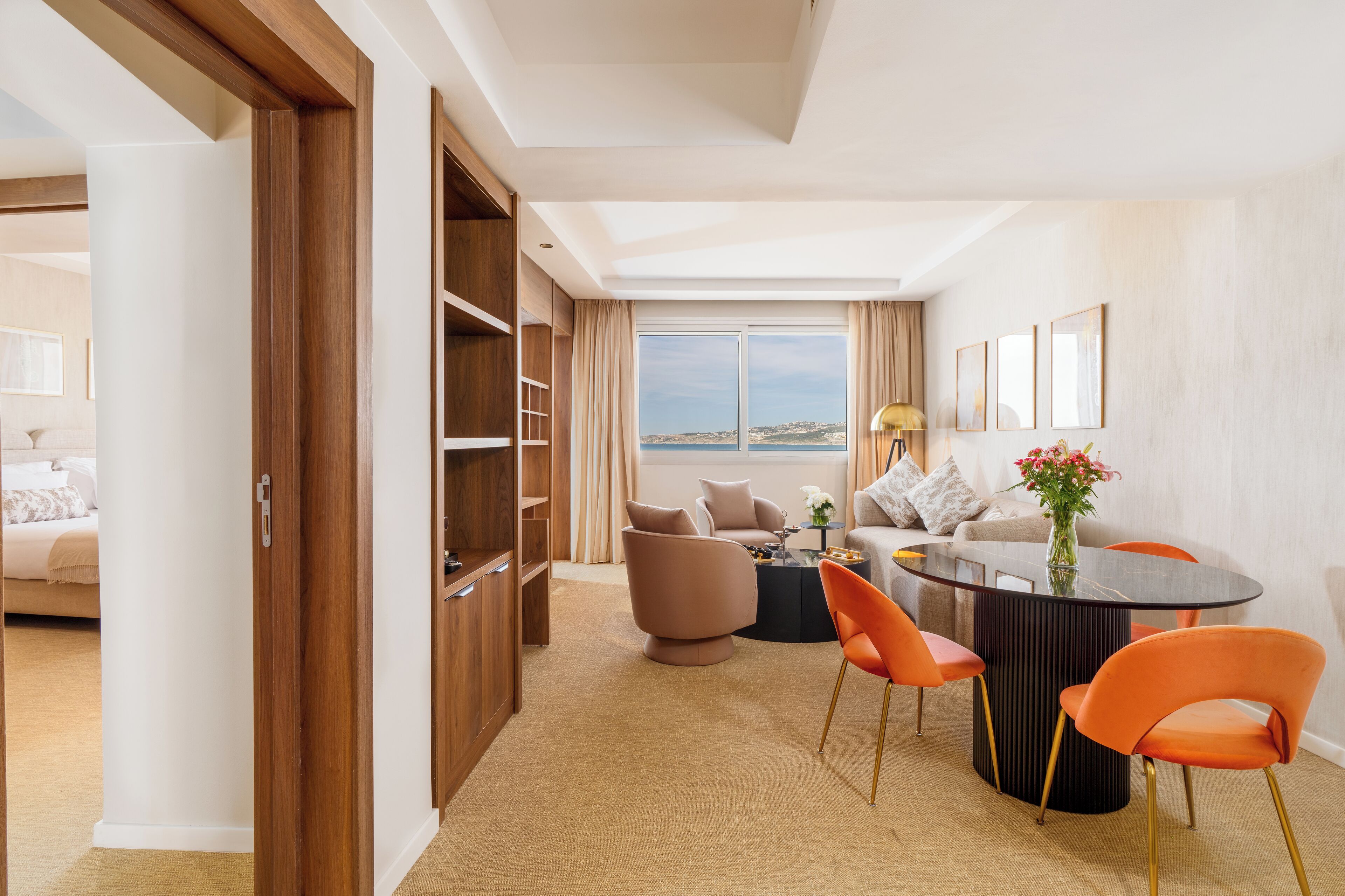 Una habitación de hotel moderna con un ventanal grande, una zona de comedor con sillas color naranja, una mesa redonda y un sofá.