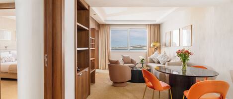 A modern hotel room with a large window, a dining area with orange chairs, a round table, and a sofa.