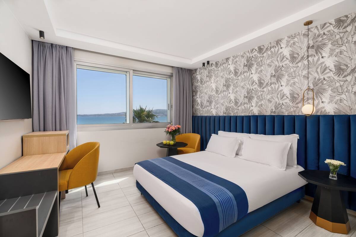 A hotel room with a large bed, a desk, a chair, a small table with a vase of flowers, and a view of the sea through a large window.
