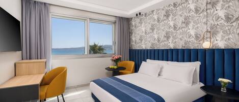 Habitación de hotel con una cama grande, un escritorio, una silla, una mesita con un jarrón de flores y vistas al mar a través de un ventanal amplio.