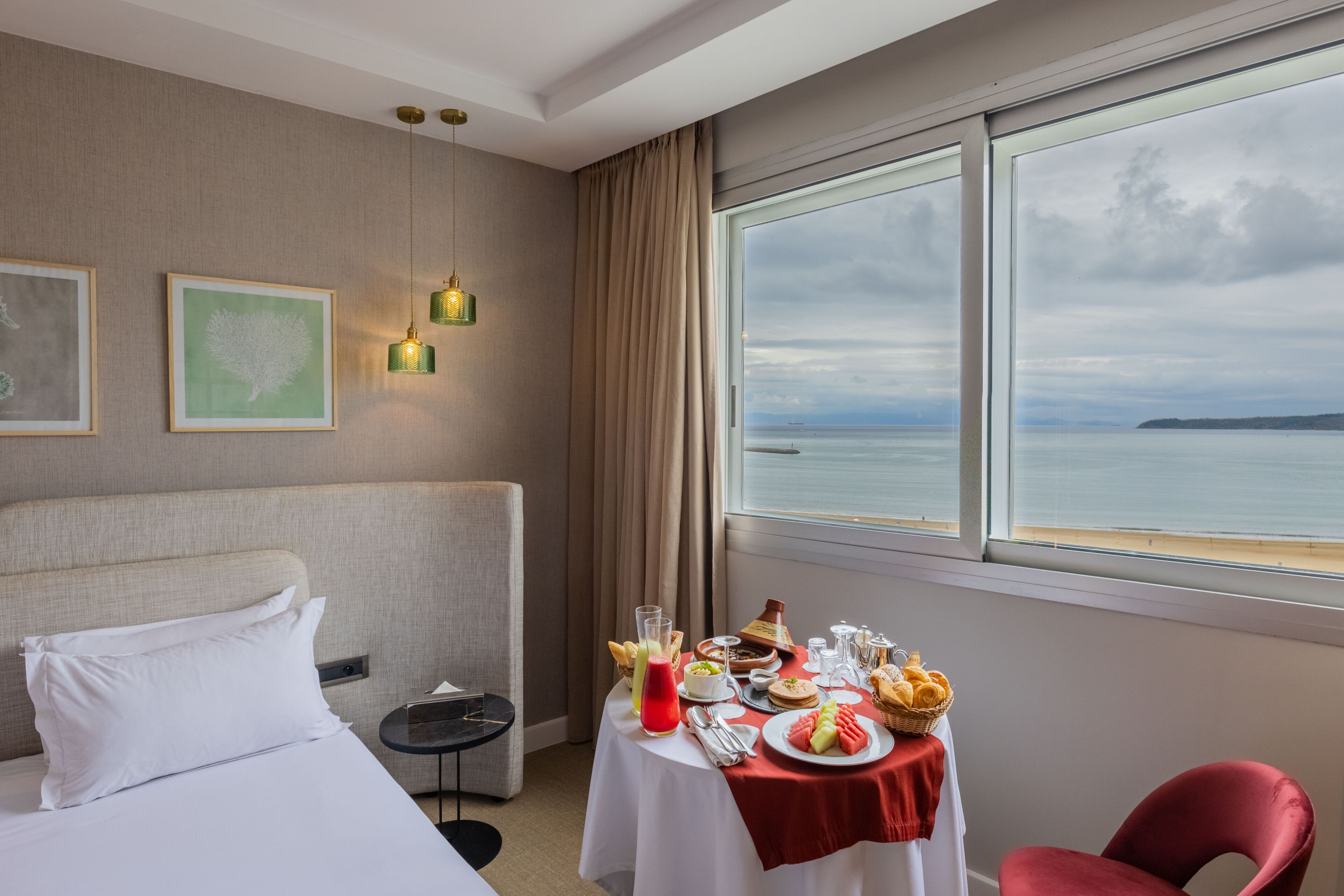 A hotel room with a bed, a table with breakfast, a chair, and a large window with a view of the sea.