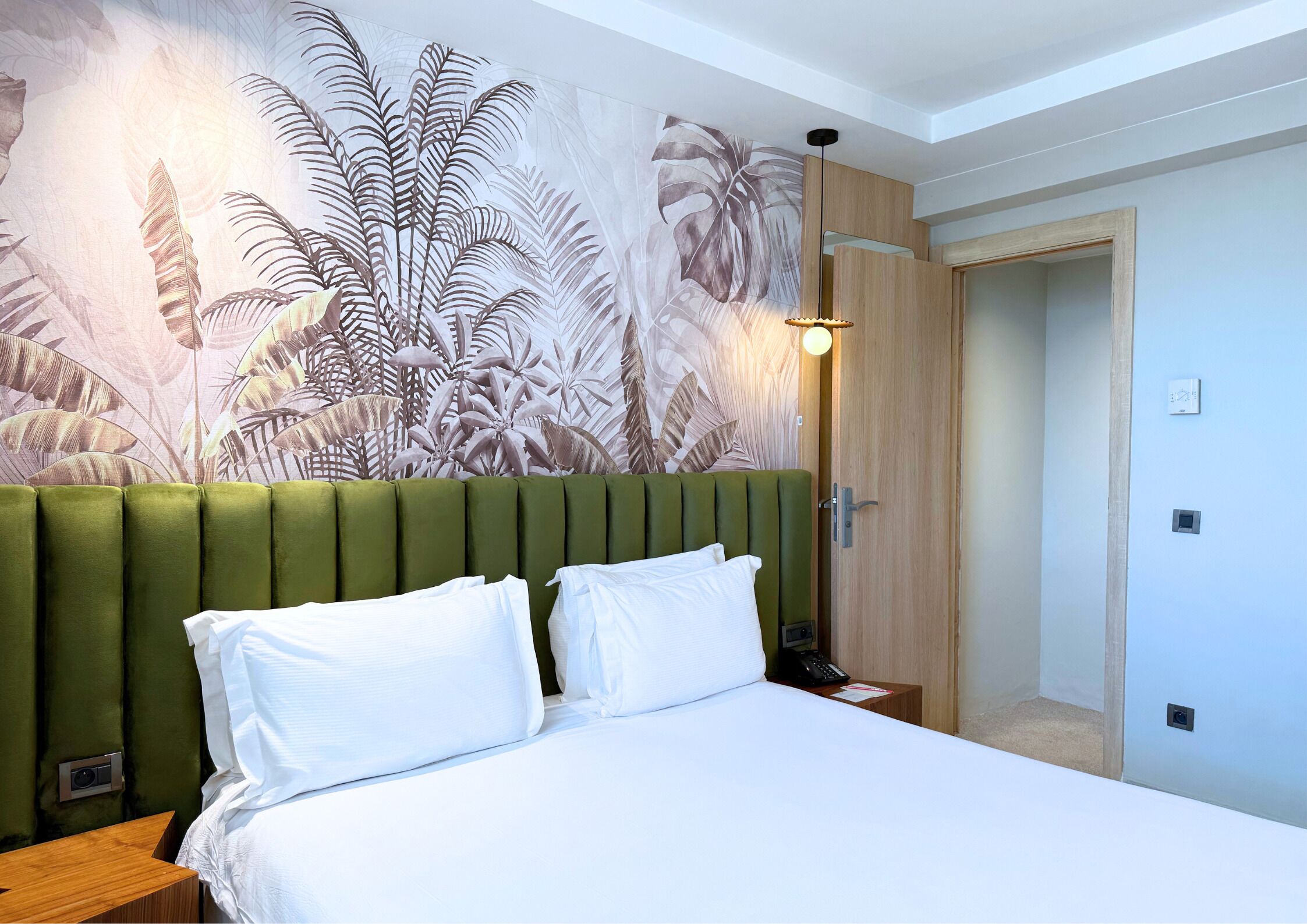 Une chambre d’hôtel avec une tête de lit recouverte d’un tissu vert, du linge de lit blanc et une grande fresque murale représentant des feuilles tropicales.