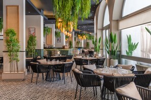 En restaurant med runde borde og sorte stole, indeplanter, hængende lys og et mønstret gulv.