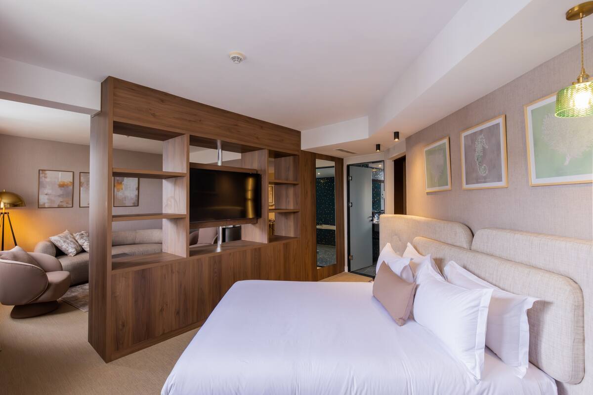 A modern hotel room with a large bed, a built-in TV, a seating area with a sofa, and a wooden partition.