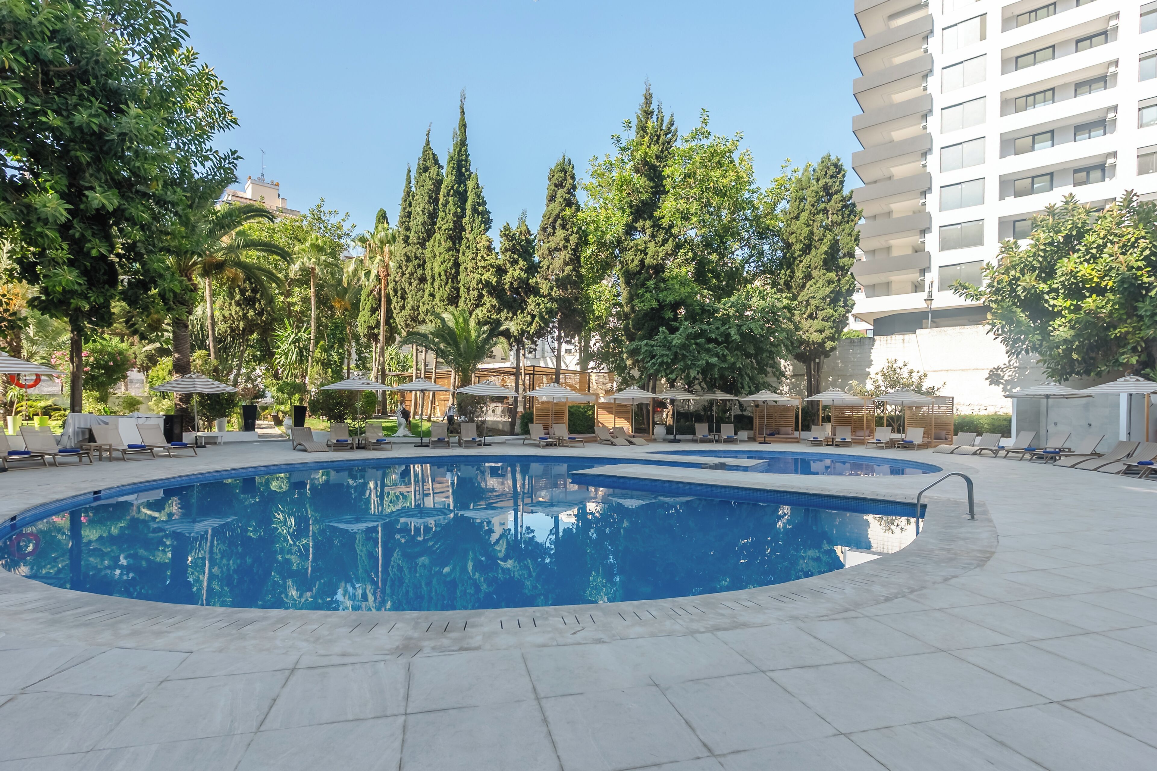 Una piscina con sillas de descanso y sombrillas, rodeada de árboles y un edificio alto.