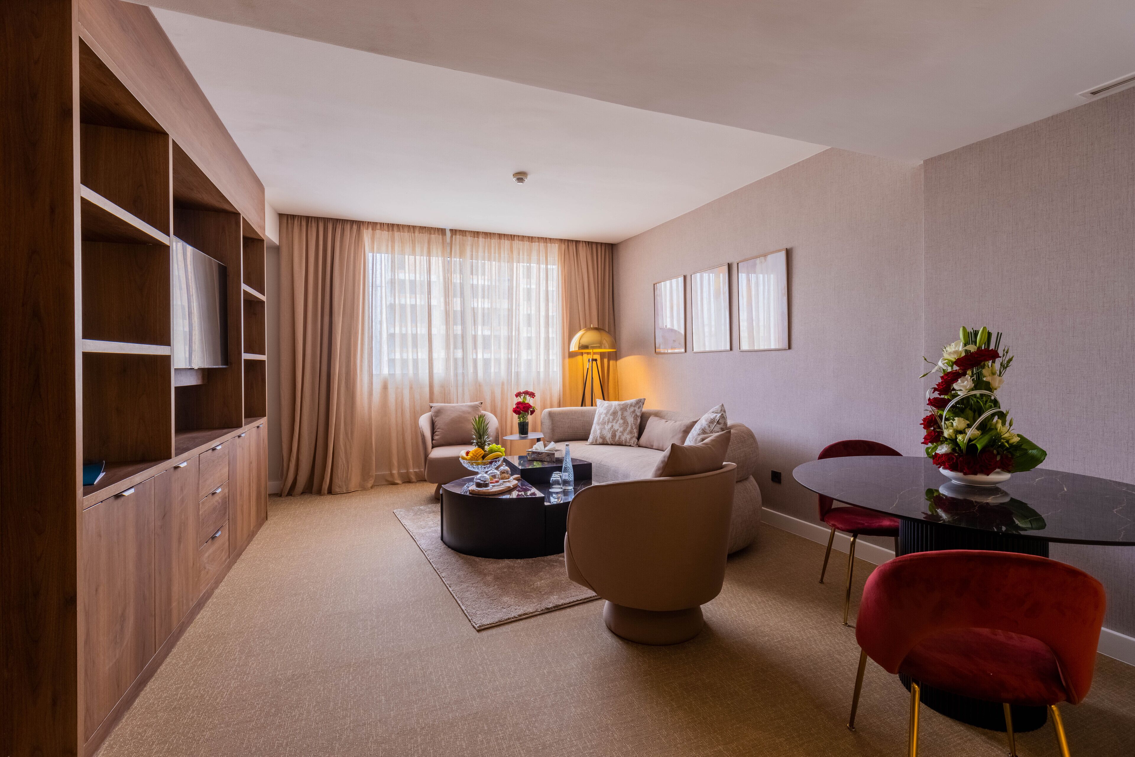 A modern living room with a sofa, coffee table, and red chairs. There is a wooden cabinet with shelves and a flat-screen TV.