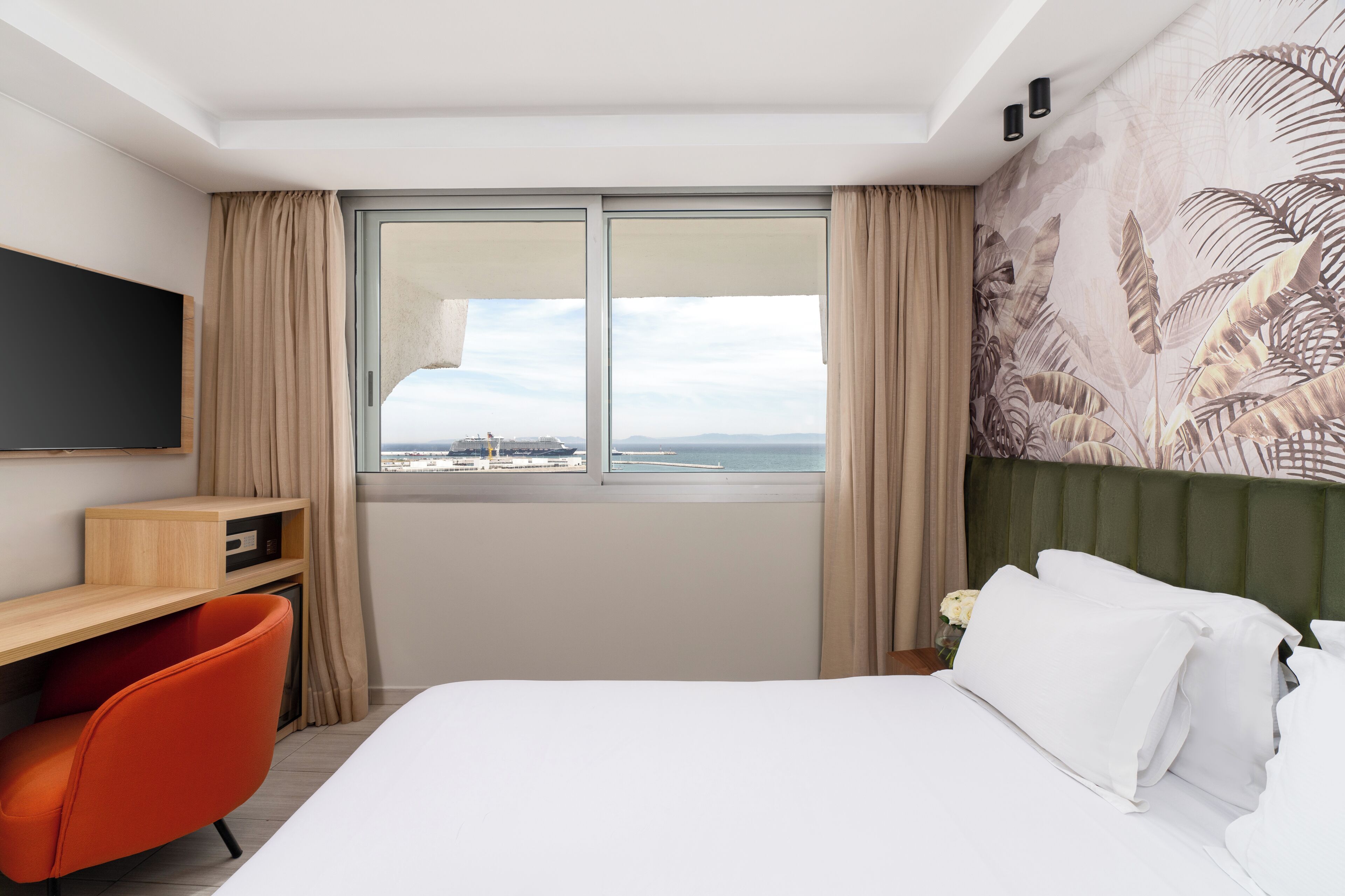 Habitación de hotel con cama, escritorio, silla, televisor y un amplio ventanal con vista al mar.
