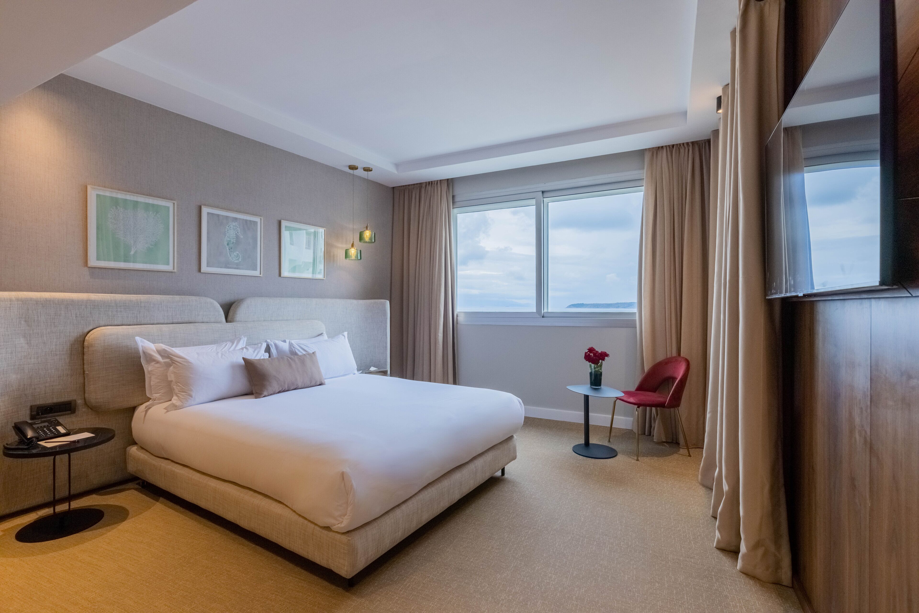 A hotel room with a large bed, a small table with a vase, a chair, and a window with curtains.