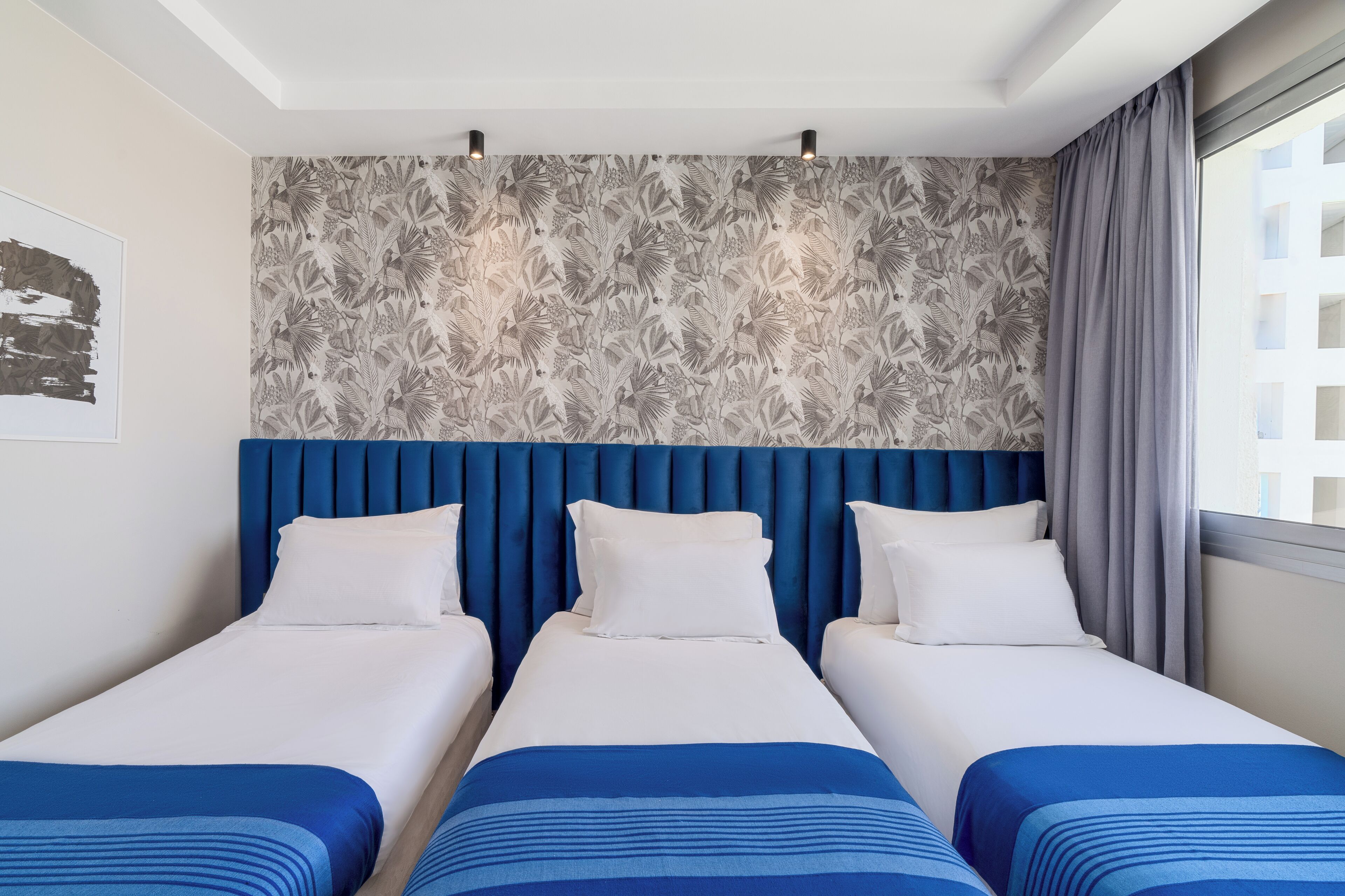 Habitación de hotel con tres camas, un papel tapiz estampado, un cabecero azul y un ventanal con cortinas.