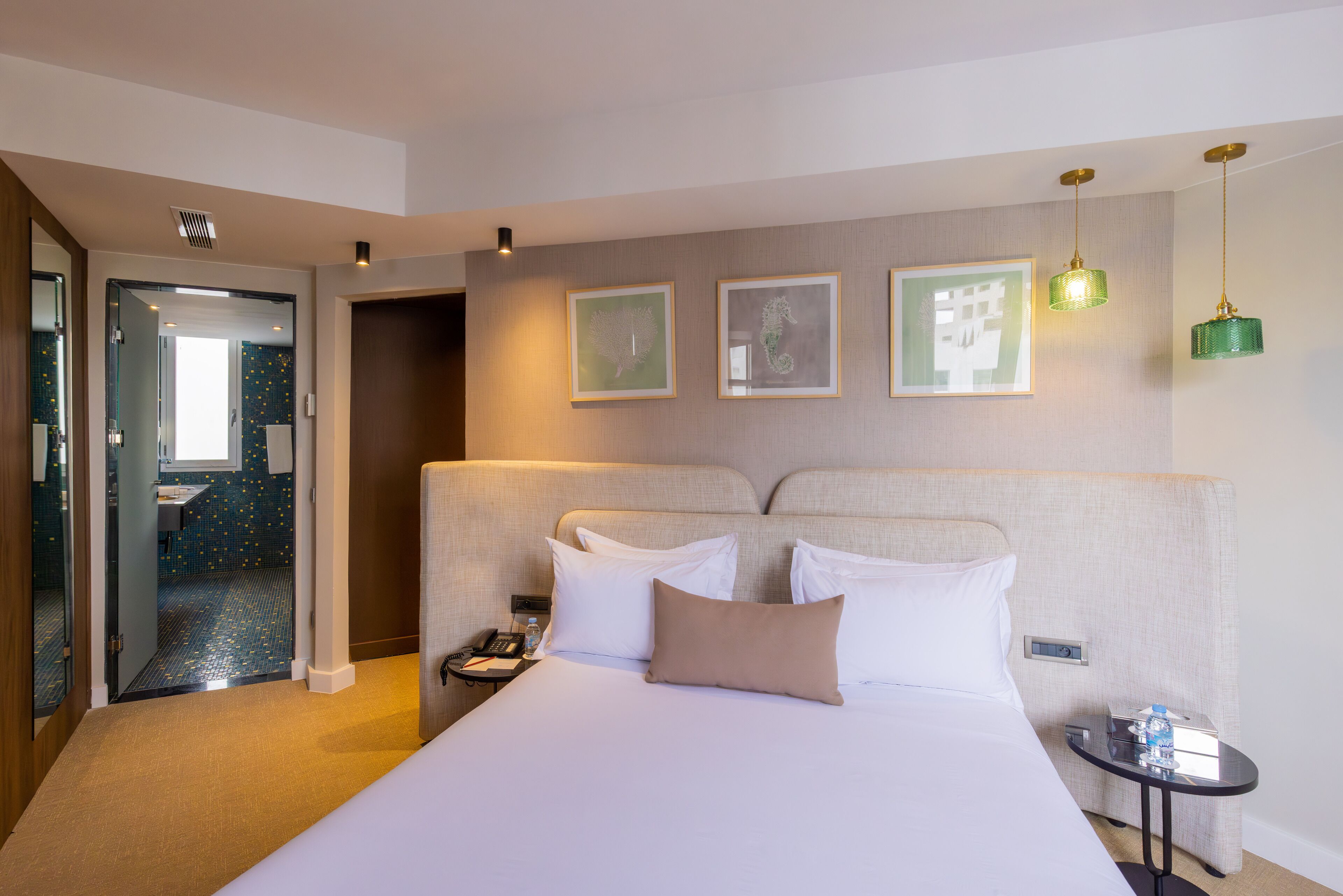 A hotel room with a large bed, a bedside table with a phone and a bottle, and a small table with a lamp. There are framed pictures on the wall and a door leading to a bathroom.