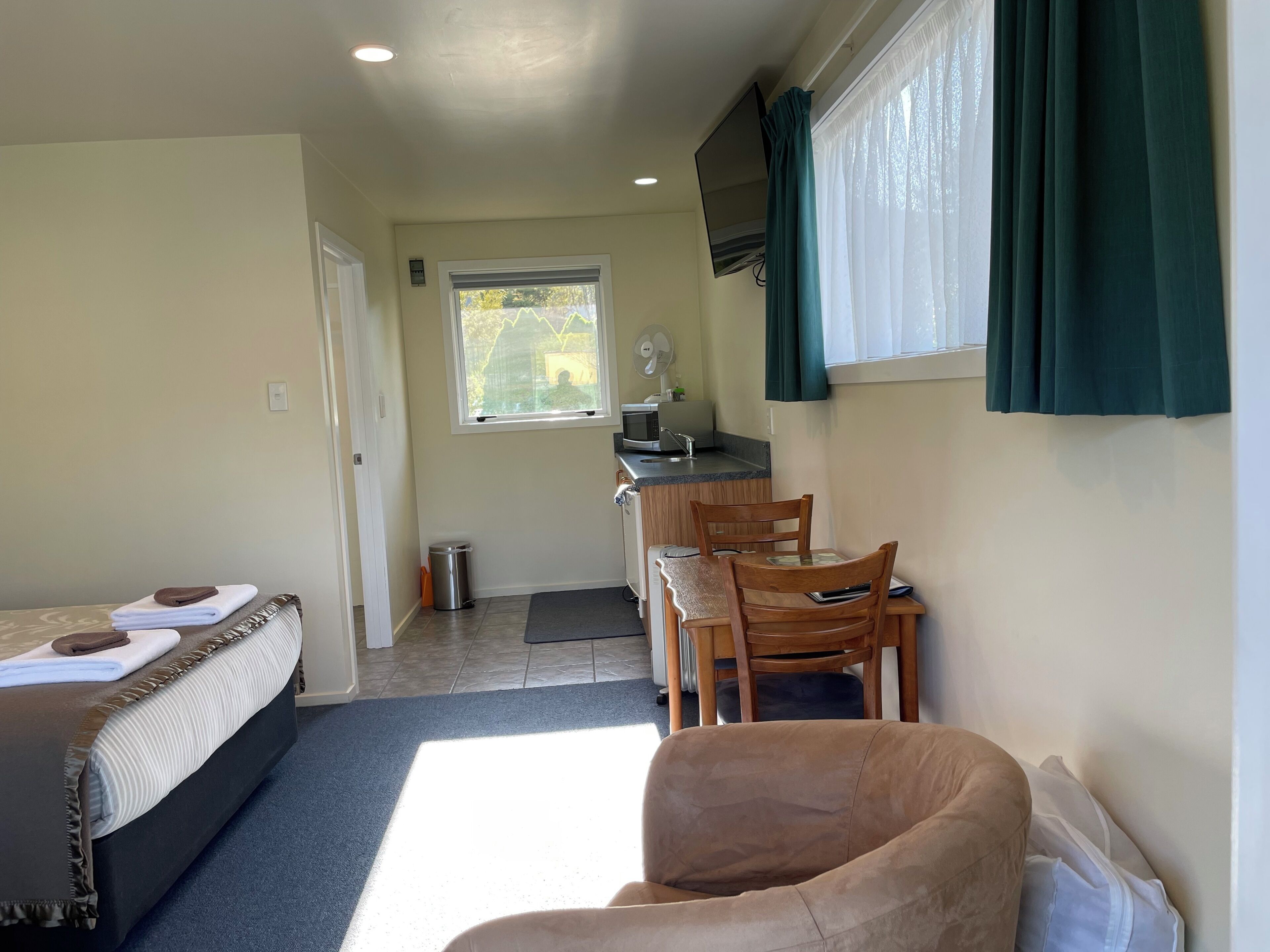 Habitación de hotel compacta con cama, kitchenette pequeña, zona de comedor con sillas y ventana con cortinas.