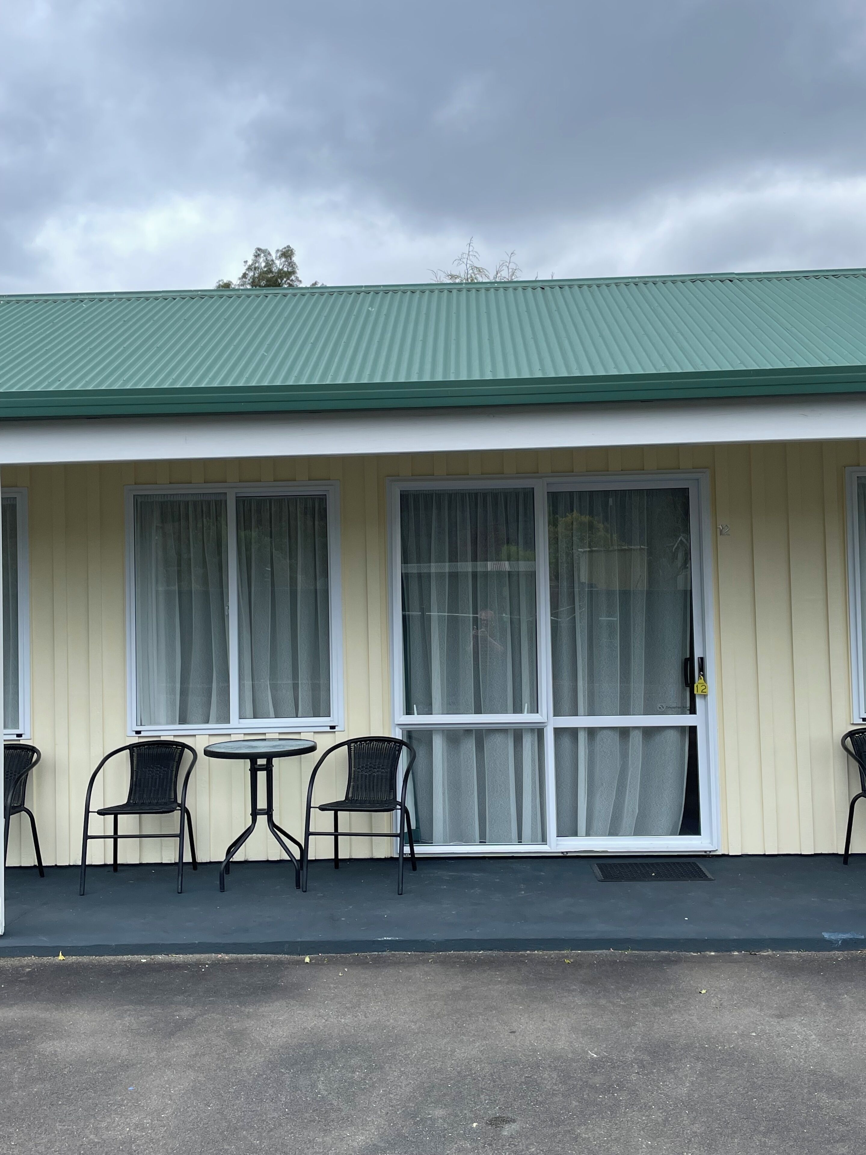 A motel with a green roof, large windows with curtains, and a small patio with chairs.