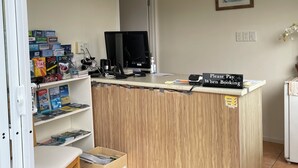 A reception desk with a sign instructing to pay when booking, a computer monitor, and a shelf with various items including books and a coffee machine. - Mataki Motels (Murchison)