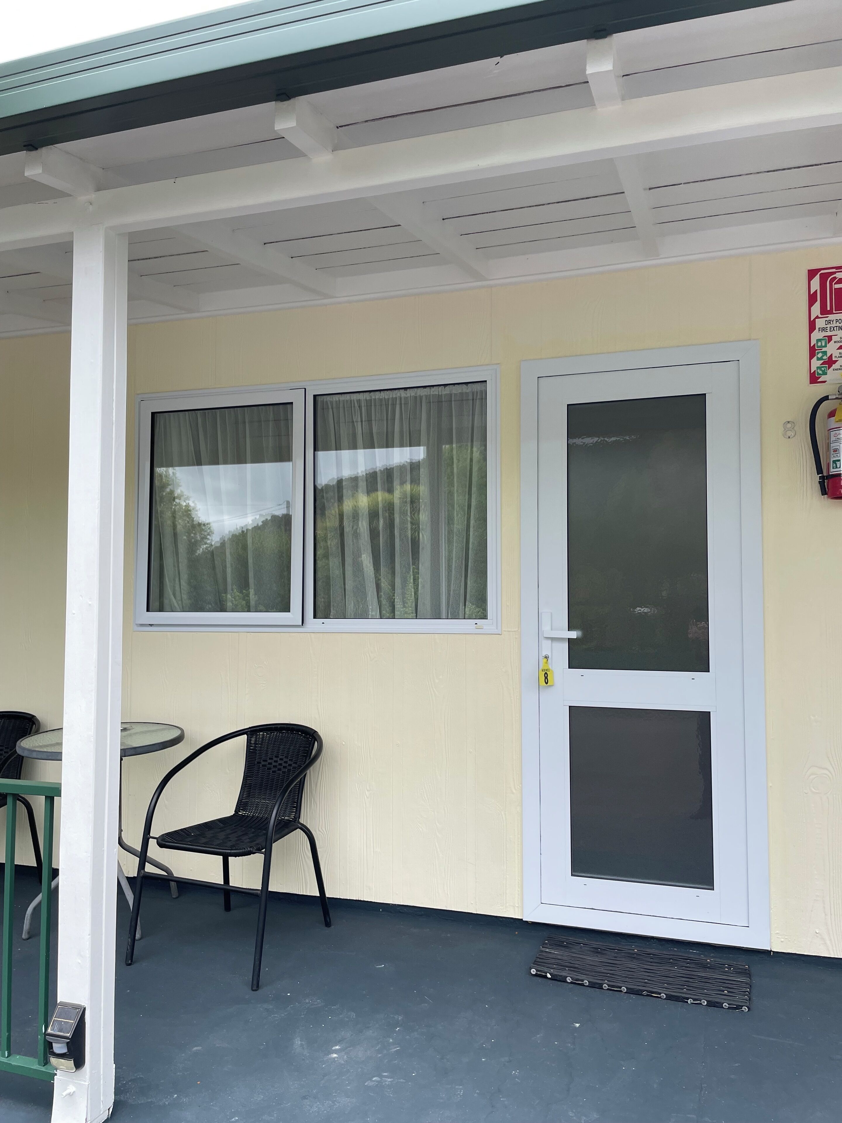 Un espacio al aire libre techado con una puerta, dos ventanas con cortinas, una silla y un extintor en la pared.