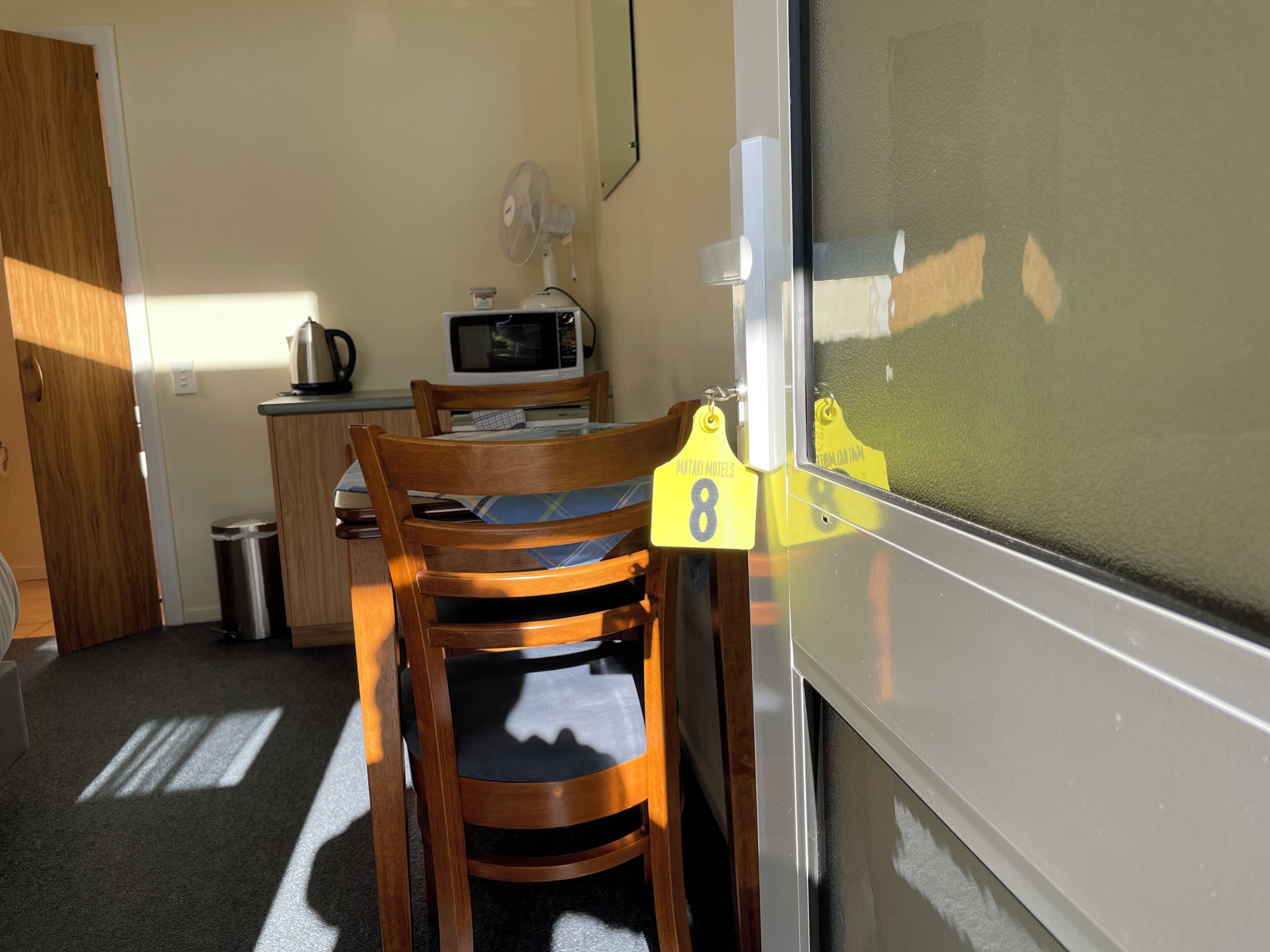 A compact kitchen with a microwave, a small trash bin, and a wooden chair with a cushion.