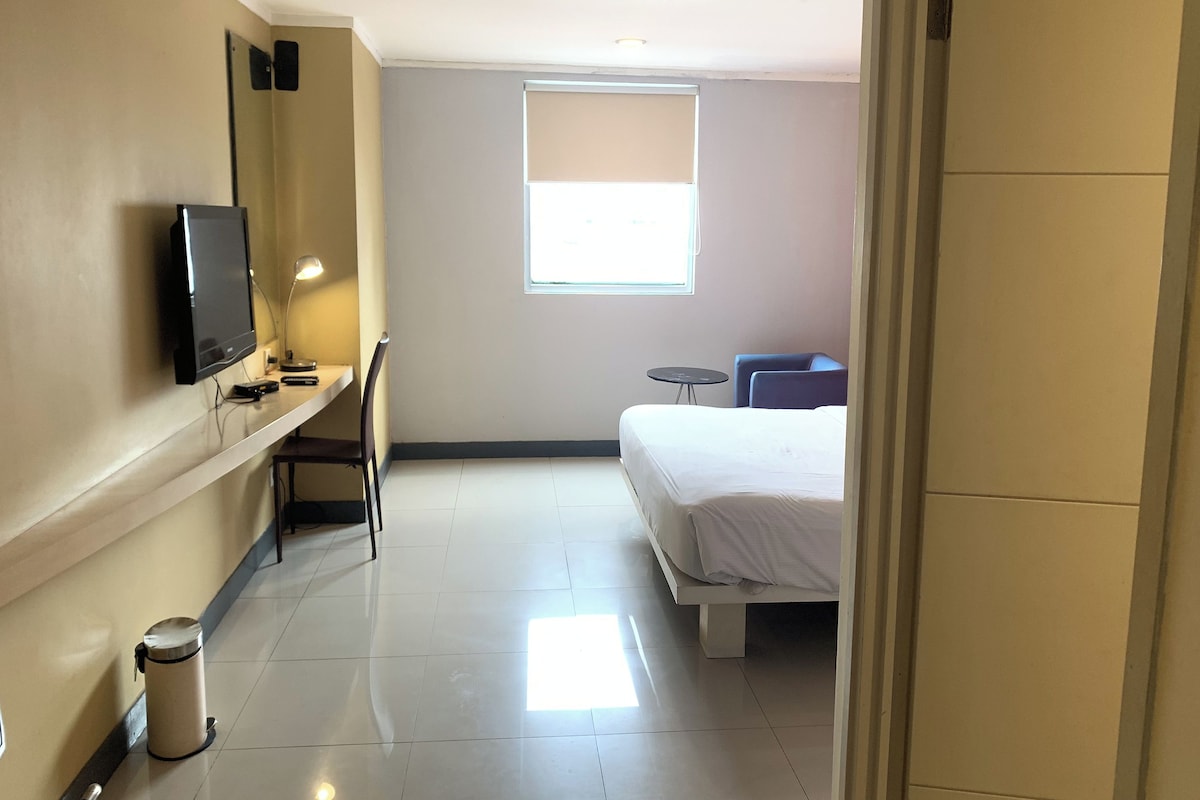 A hotel room with a bed, a desk with a computer, a chair, a small table, a TV, and a window.