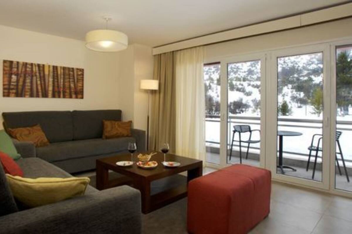 Een moderne woonkamer met een grijze bank, een houten salontafel met wijnglazen, en een rode poef. Er is een groot raam met uitzicht op bomen en een balkon met buitenmeubilair.