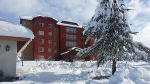 A multi-story building with a red facade and multiple windows, surrounded by snow-covered ground and trees. - Village Condo (San Carlos de Bariloche)