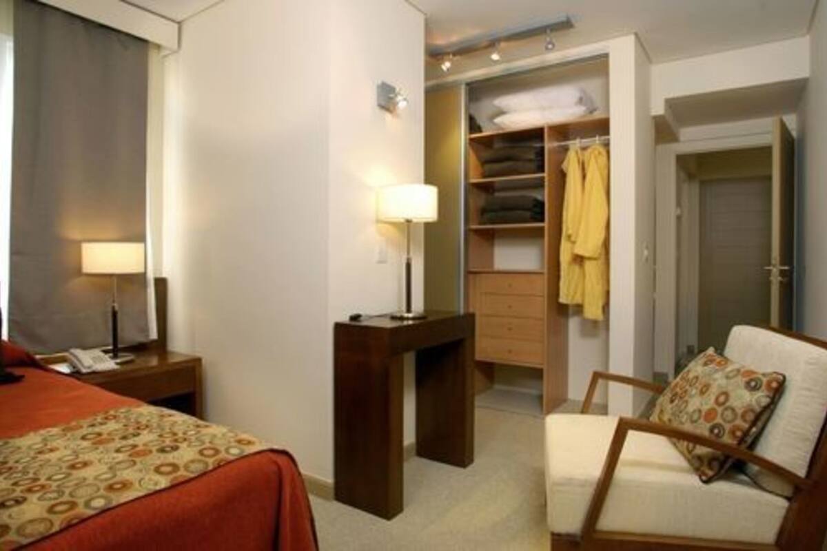 Een hotelkamer met een bed, nachtkastje met lamp, stoel met kussen, garderobe en bureau met lamp.