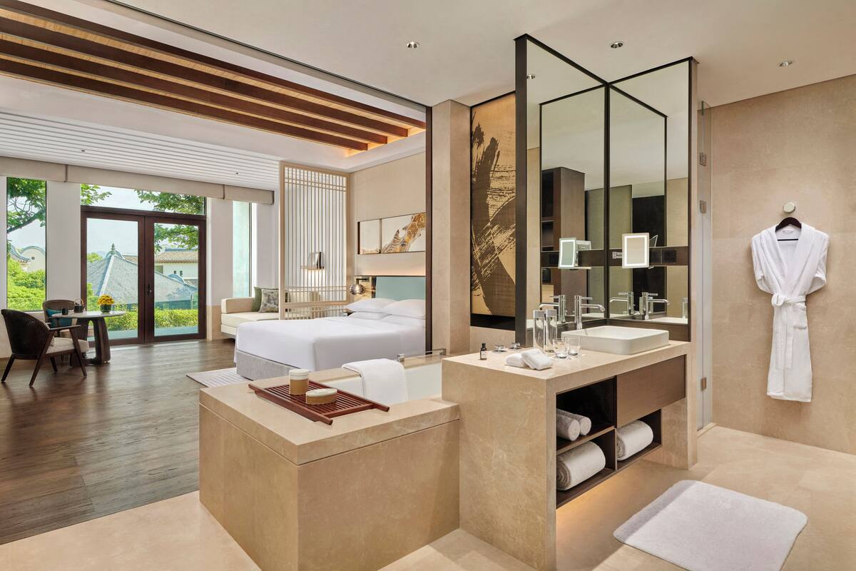 Une chambre d’hôtel moderne dotée d’un grand lit, d’un coin salon avec une table et des chaises, d’une salle de bain avec un grand miroir et d’une vue sur la verdure environnante.