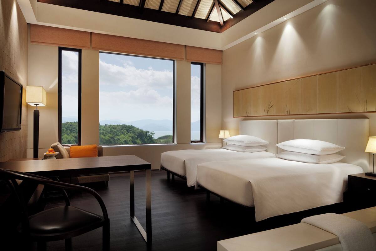 Une chambre d’hôtel avec deux lits, un bureau, une chaise et de grandes fenêtres donnant sur l’extérieur.