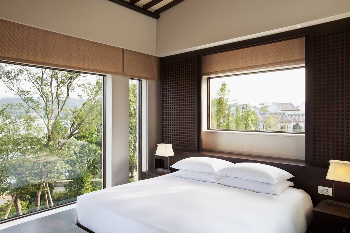 Une chambre d’hôtel moderne, dotée d’un grand lit, de deux tables de chevet et offrant une vue sur les arbres et les bâtiments grâce à de grandes fenêtres.