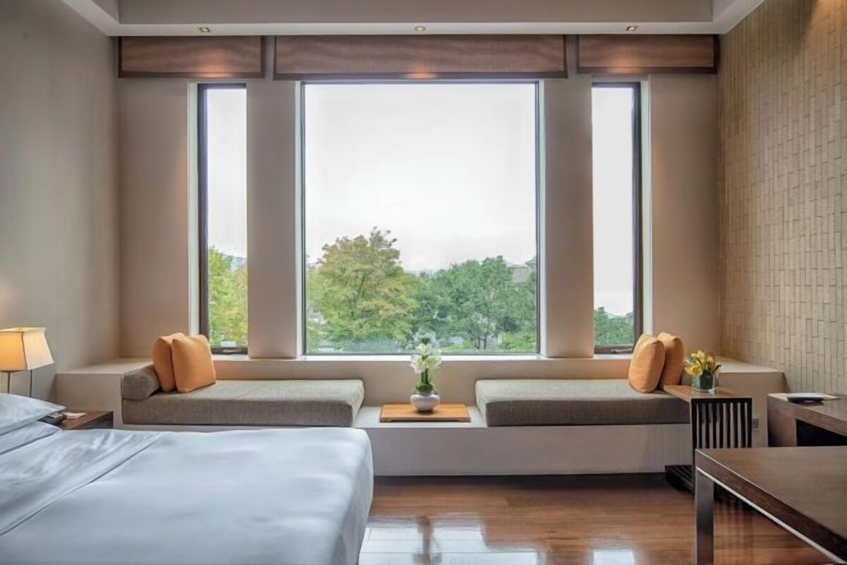 Quarto de hotel moderno com uma janela grande, uma cama, um espaço de estar com almofadas e uma pequena mesa com um vaso de flores.