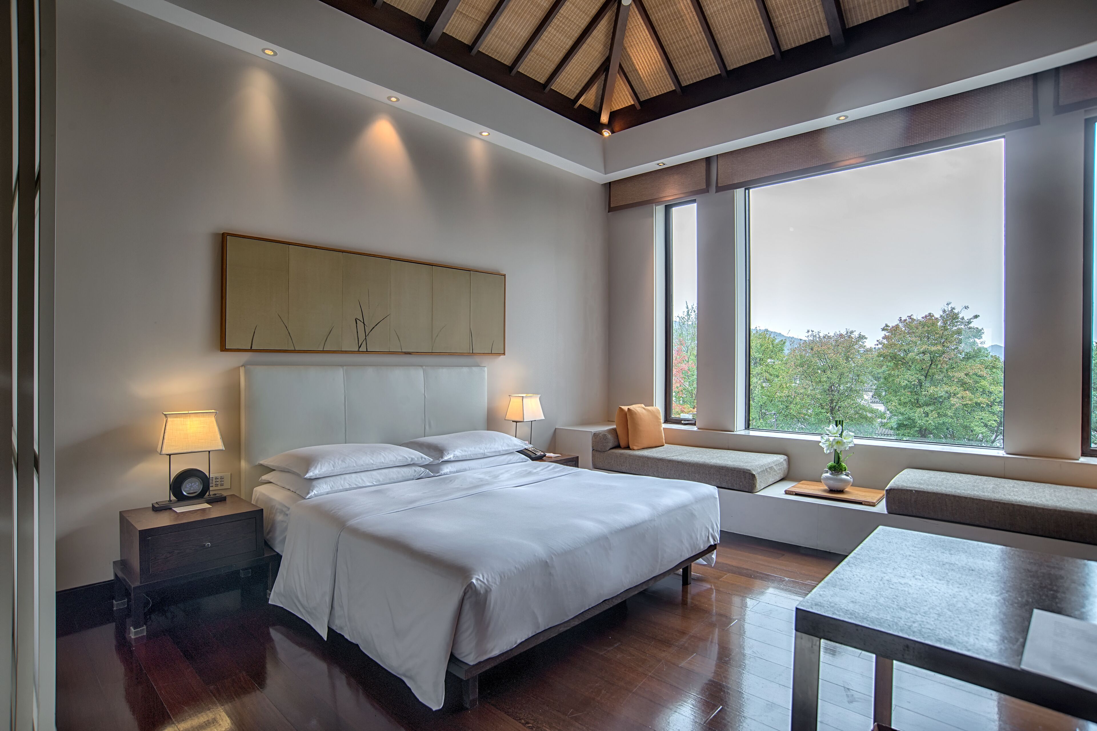 A modern bedroom with a large bed, bedside tables with lamps, a sofa, and a view of trees through large windows.