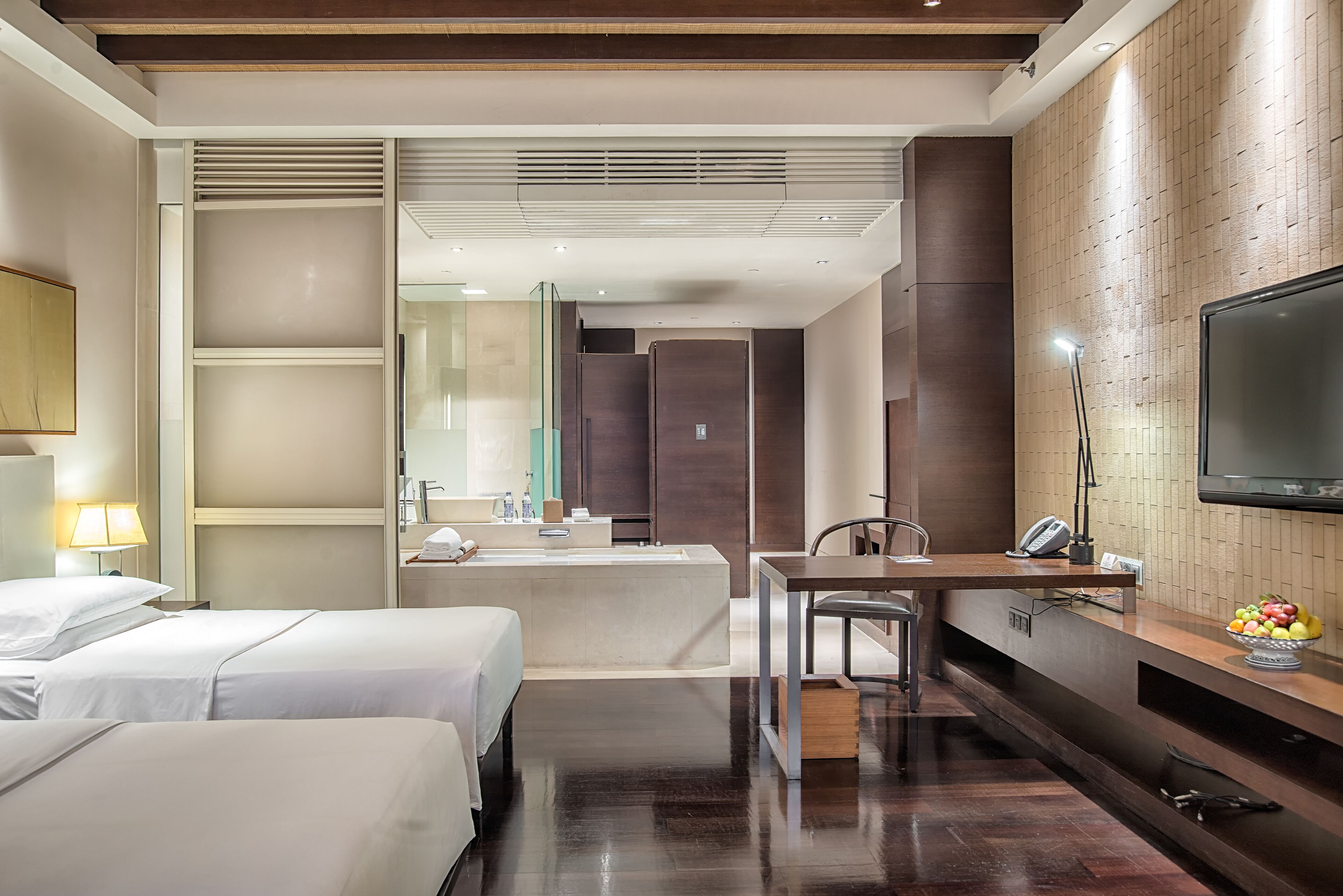 A hotel room with two beds, a desk, a TV, and a bathroom with a large mirror and sink.