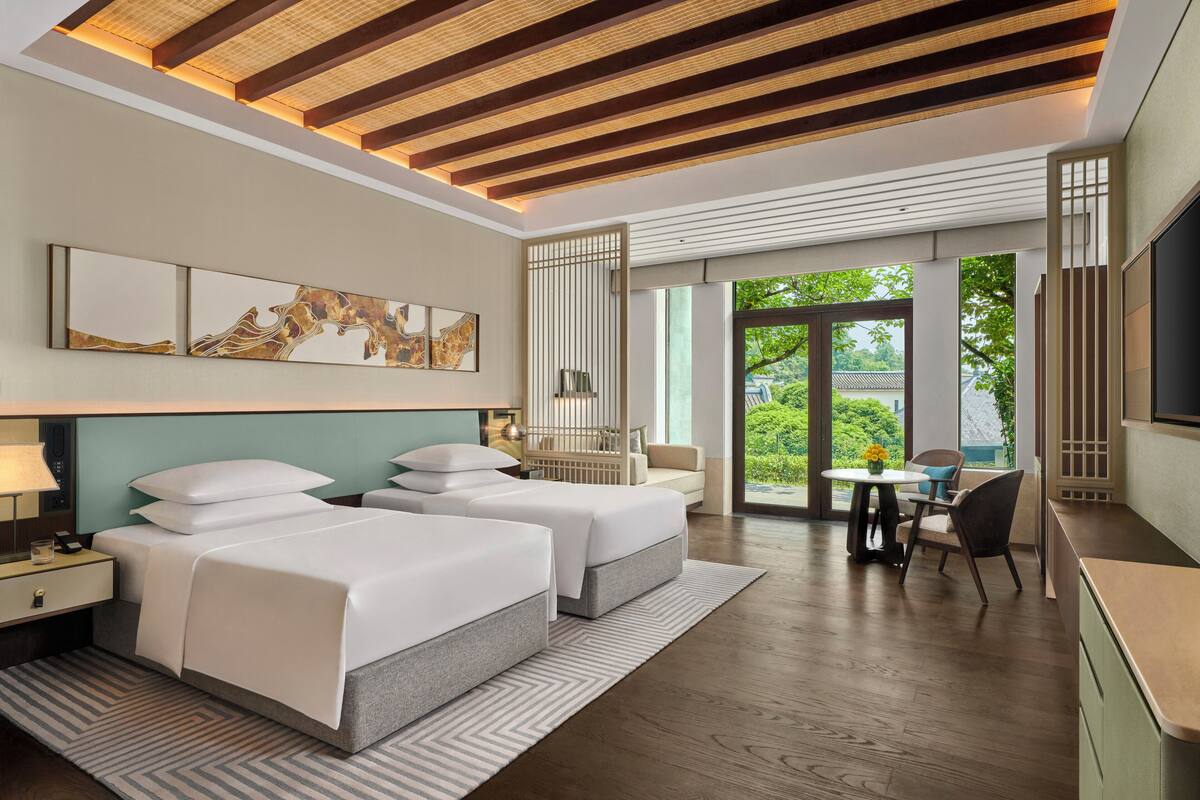 Une chambre d’hôtel moderne, dotée de deux lits, d’un bureau, d’une chaise et d’une grande fenêtre donnant sur un paysage verdoyant.