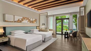 A modern hotel room with two beds, a desk, a chair, and a large window offering a view of greenery.