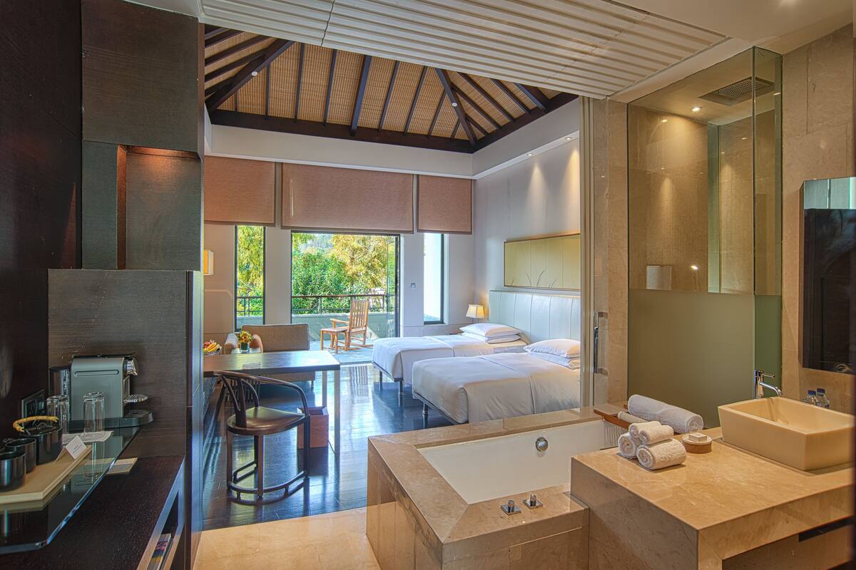 Une chambre d’hôtel moderne dotée d’un grand lit, d’une salle de bain séparée avec baignoire, d’un coin repas avec une table et des chaises, et offrant une vue sur l’extérieur grâce à de grandes fenêtres.