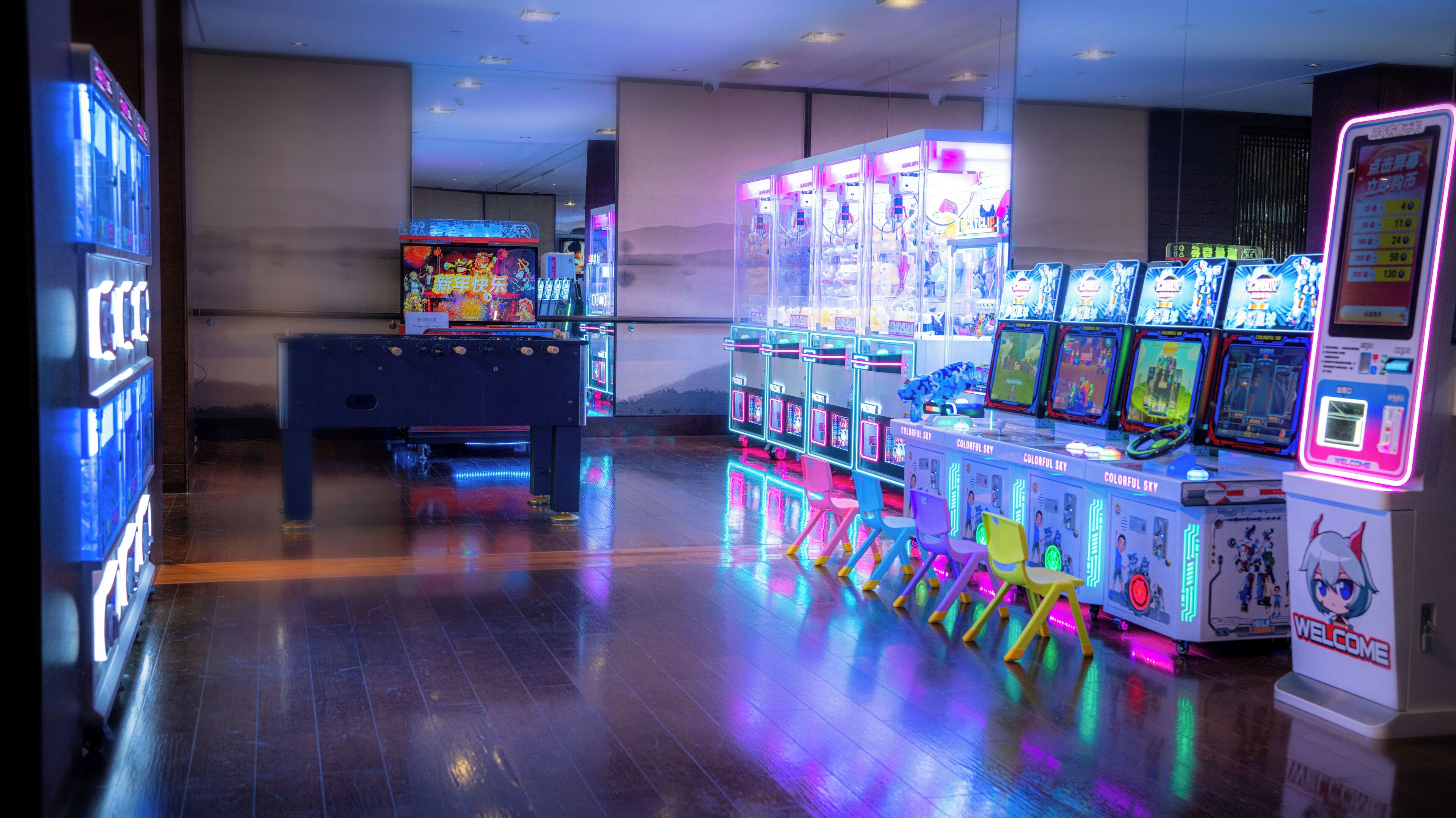 A row of arcade machines with neon lights, a pool table, and a few chairs in a room with a polished wooden floor.