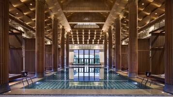 A spacious indoor swimming pool with a modern design, surrounded by wooden paneling and columns, and a view of the outdoors through large windows.
