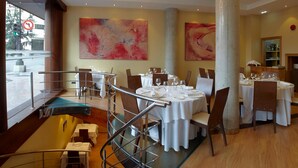 Un restaurante con mesas redondas preparadas para comer, una escalera con barandillas metálicas y pinturas abstractas en las paredes.